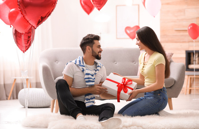 Lovely young couple with gift box in living room decorated with heart shaped balloons. Valentine's day celebration