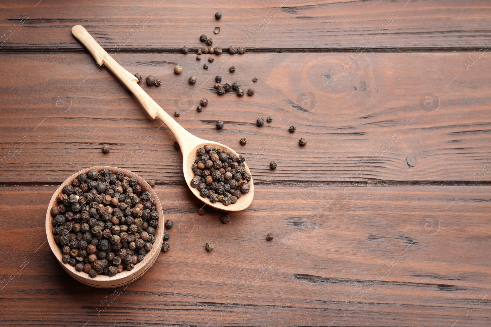 Photo of Bowl and spoon of black pepper corns on wooden table, flat lay with space for text