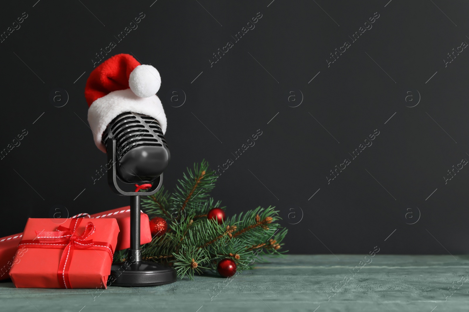 Photo of Retro microphone with Santa hat, gift boxes and festive decor on wooden table against black background, space for text. Christmas music