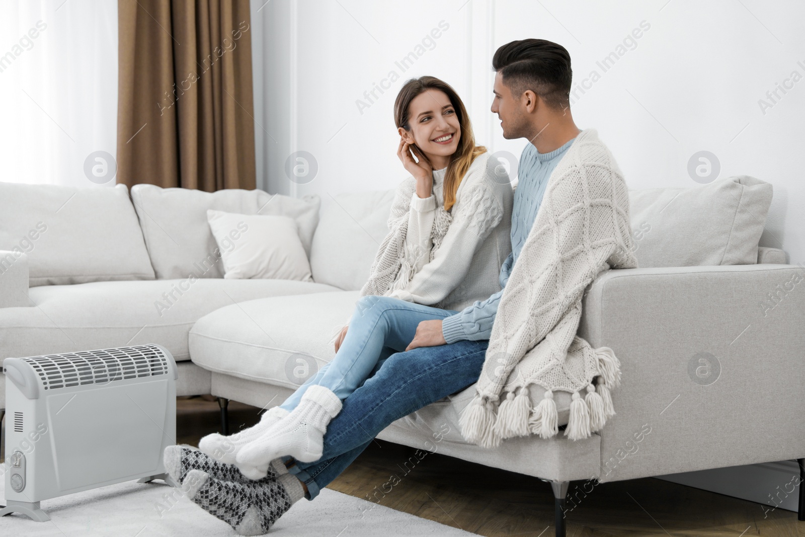 Photo of Happy couple sitting on sofa near electric heater at home