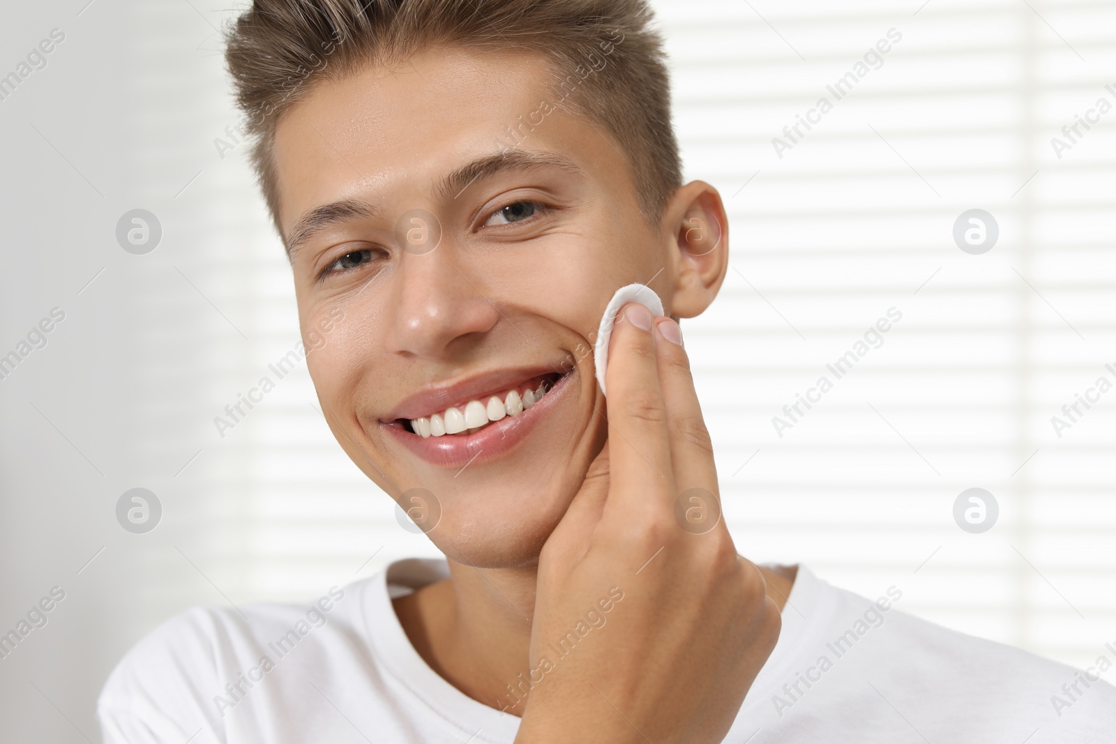 Photo of Handsome young man cleaning face with cotton pad indoors