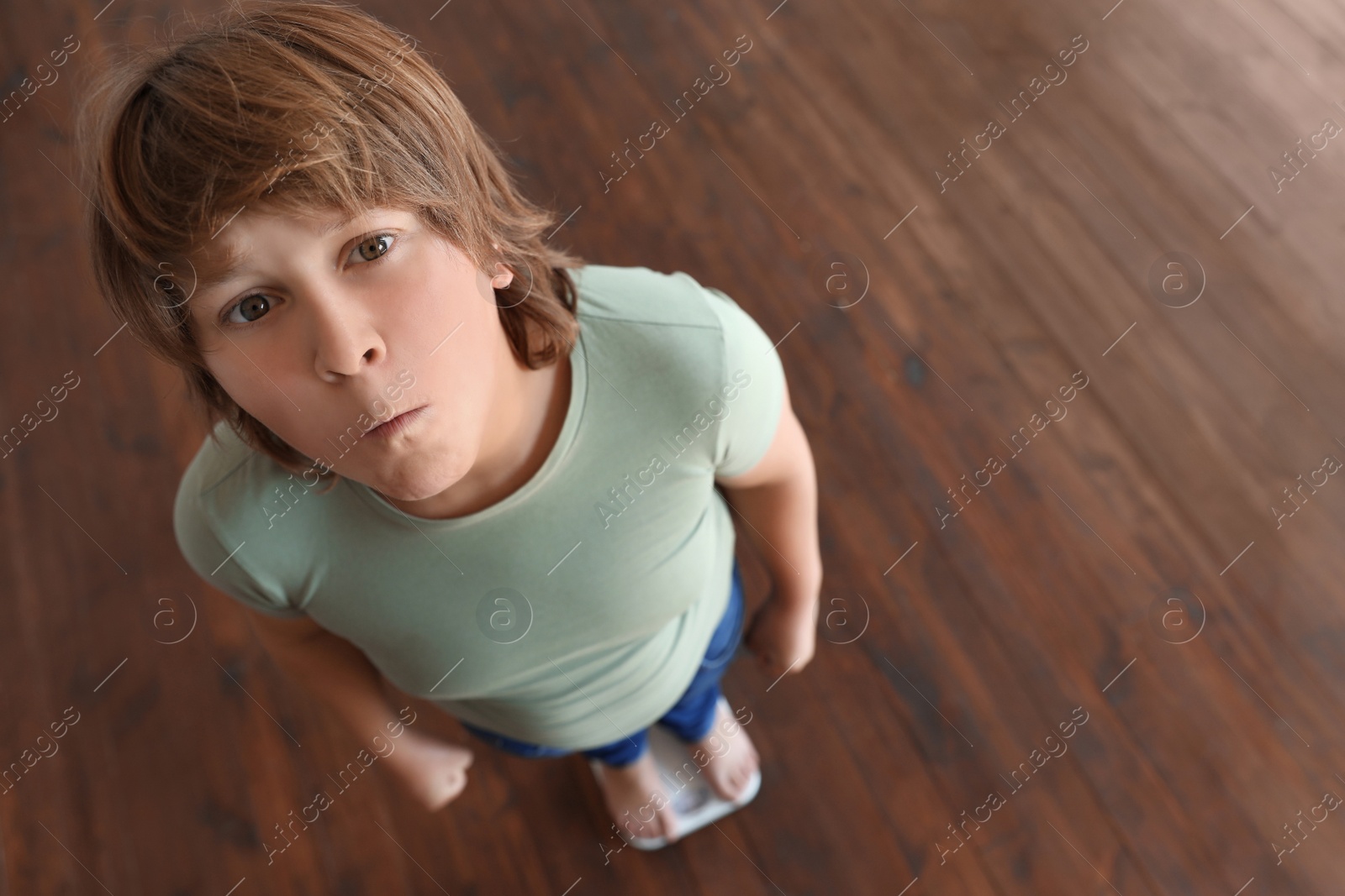 Photo of Emotional overweight boy standing on floor scales indoors, above view. Space for text