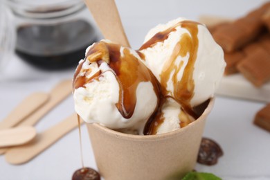 Photo of Scoops of ice cream with caramel sauce in paper cup on white table, closeup