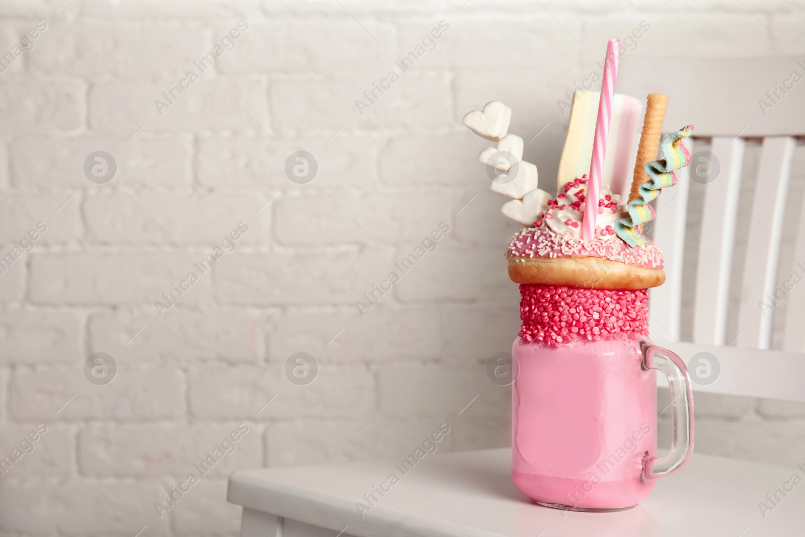 Photo of Tasty milk shake with sweets in mason jar on chair near brick wall. Space for text