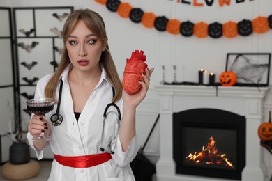 Woman in scary nurse costume with glass of wine and heart model indoors, space for text. Halloween celebration