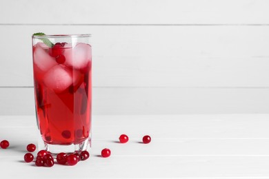 Tasty cranberry juice with ice cubes in glass and fresh berries on white wooden table. Space for text