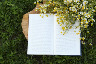 Photo of Open book and chamomiles on wooden stump outdoors, flat lay
