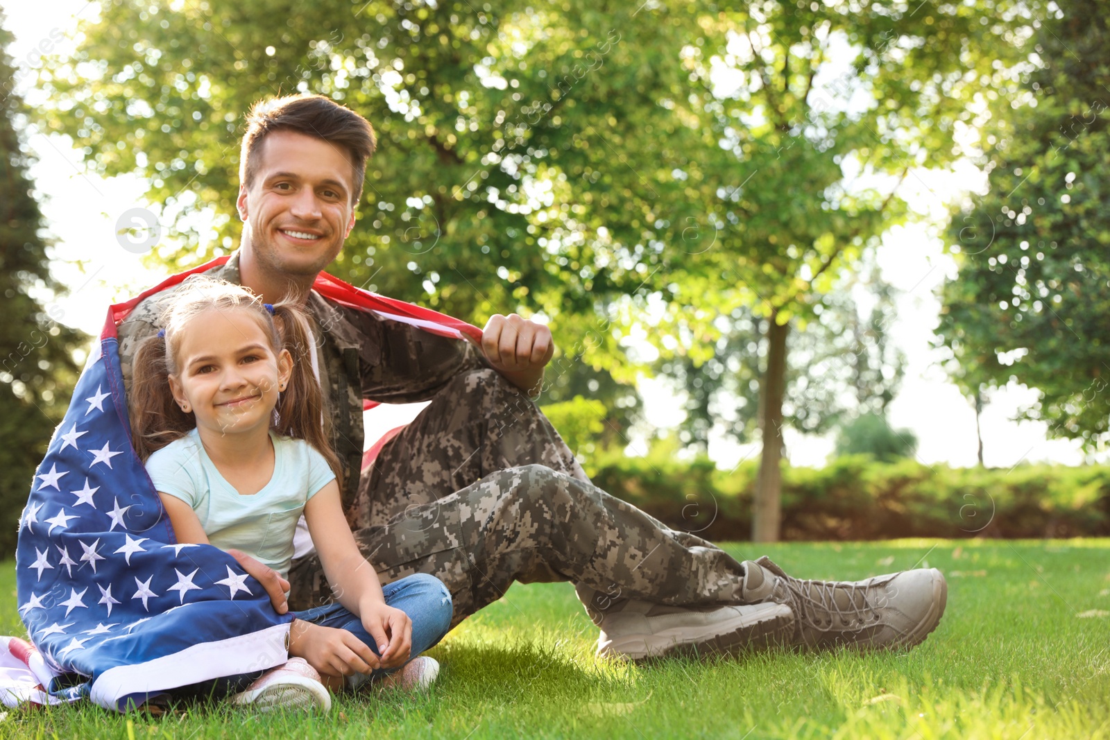 Photo of Father in military uniform with American flag and his little daughter sitting on grass at park