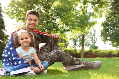 Father in military uniform with American flag and his little daughter sitting on grass at park