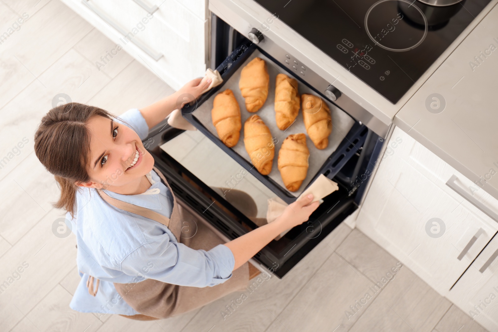 Photo of Woman taking baking tray with croissants out of oven in kitchen