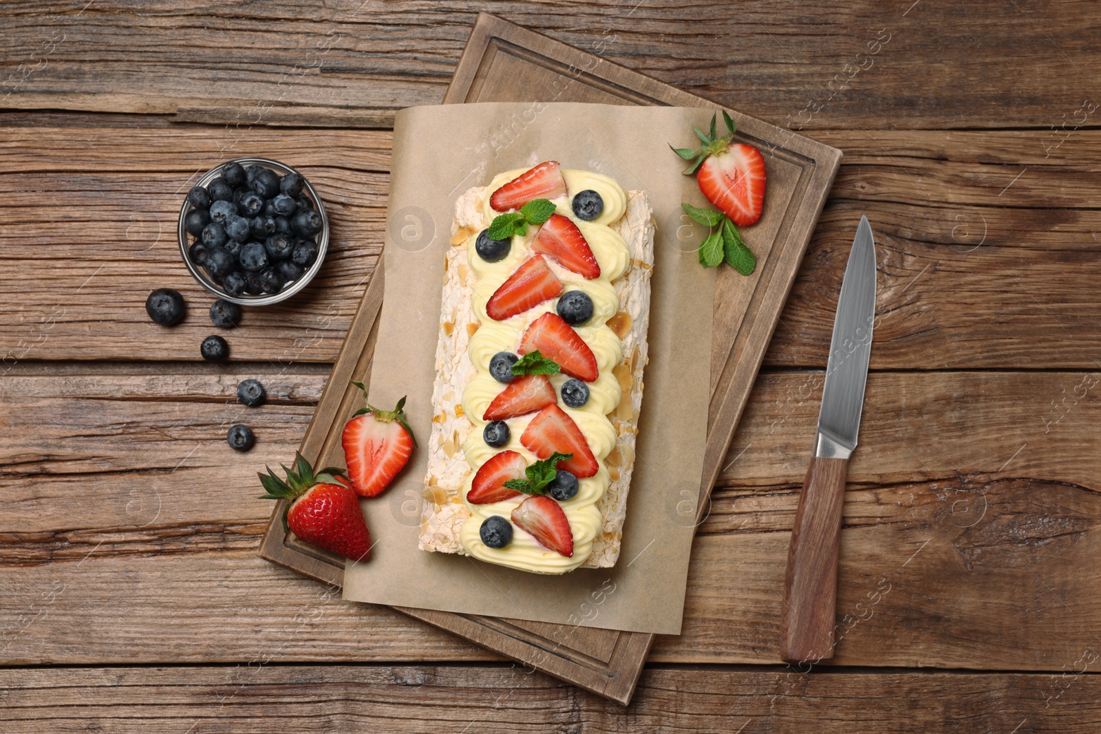 Photo of Tasty meringue roll with cream, strawberry, blueberry, mint and knife on wooden table, flat lay