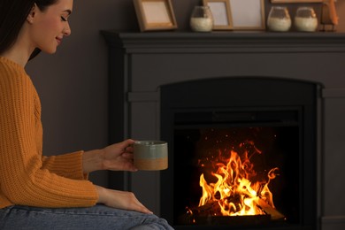 Beautiful young woman with cup of hot drink near fireplace at home. Space for text