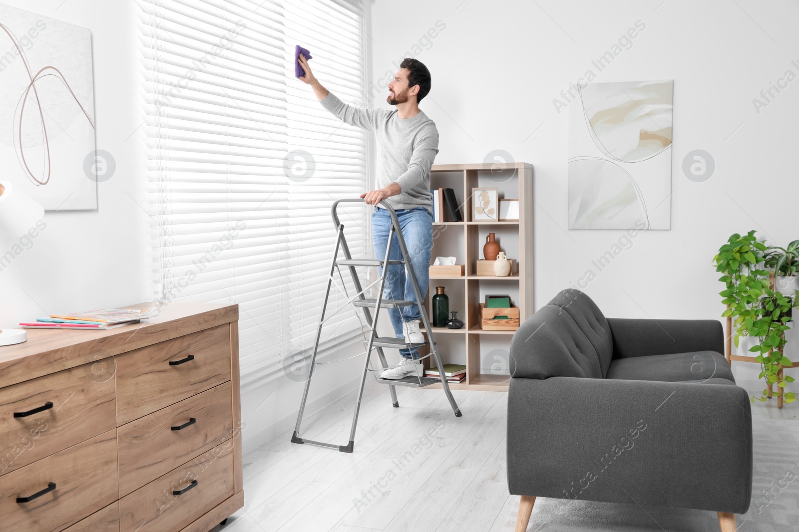 Photo of Man on metal ladder wiping blinds at home
