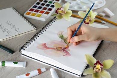 Woman drawing beautiful orchid flowers in sketchbook at wooden table, closeup