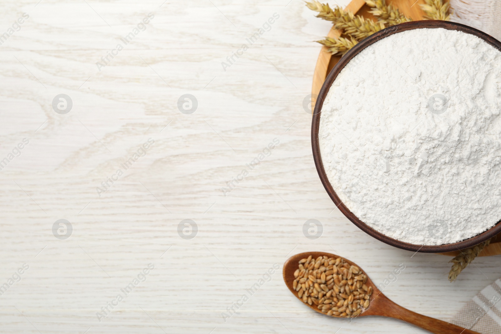 Photo of Organic flour in bowl, spoon with grains of wheat and spikelets on white wooden table, flat lay. Space for text