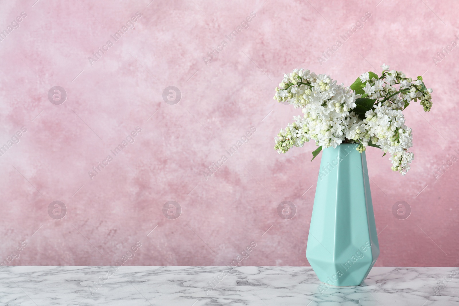 Photo of Blossoming lilac flowers in vase on marble table against color background. Space for text