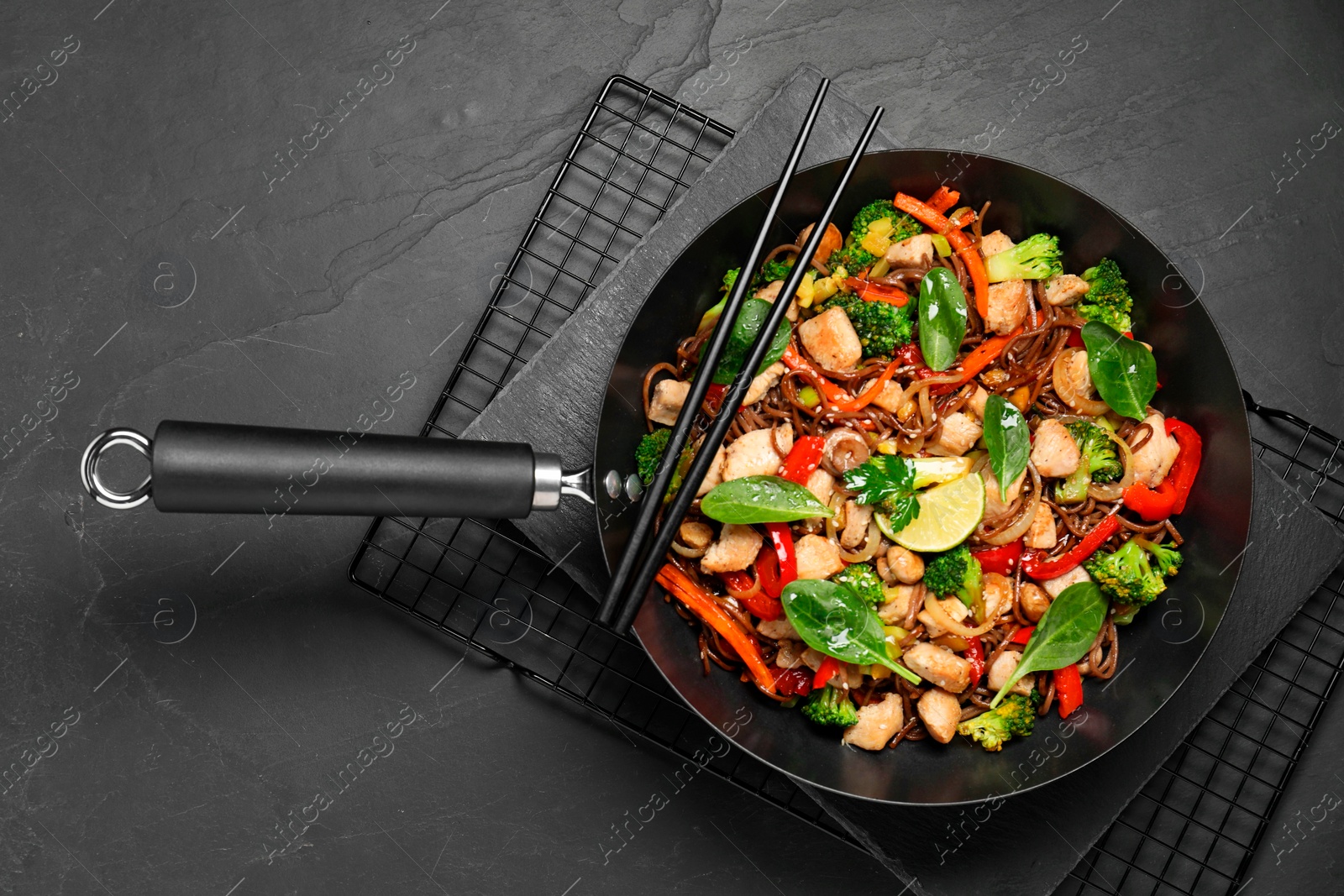 Photo of Stir-fry. Tasty noodles with meat in wok and chopsticks on grey textured table, top view