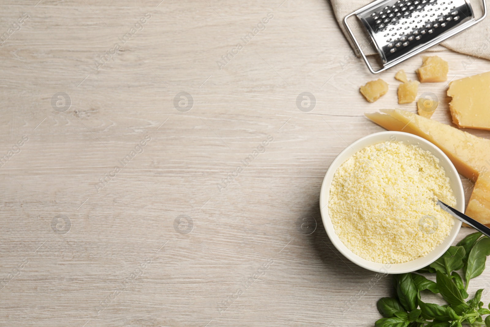 Photo of Delicious grated parmesan cheese in bowl, grater and basil on white wooden table, flat lay. Space for text