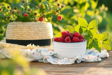 Photo of Tasty ripe raspberries in bowl, green leaves and straw hat on wooden table outdoors