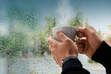 Image of Man with cup of hot drink near window on rainy day, closeup