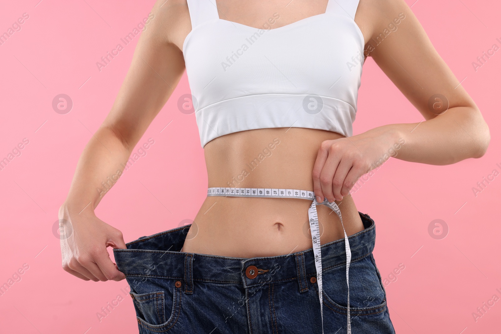 Photo of Slim woman wearing big jeans and measuring waist with tape on pink background, closeup. Weight loss