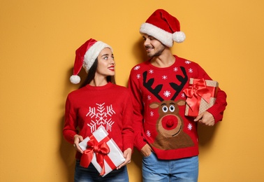 Young couple in Christmas sweaters and hats with gifts on color background