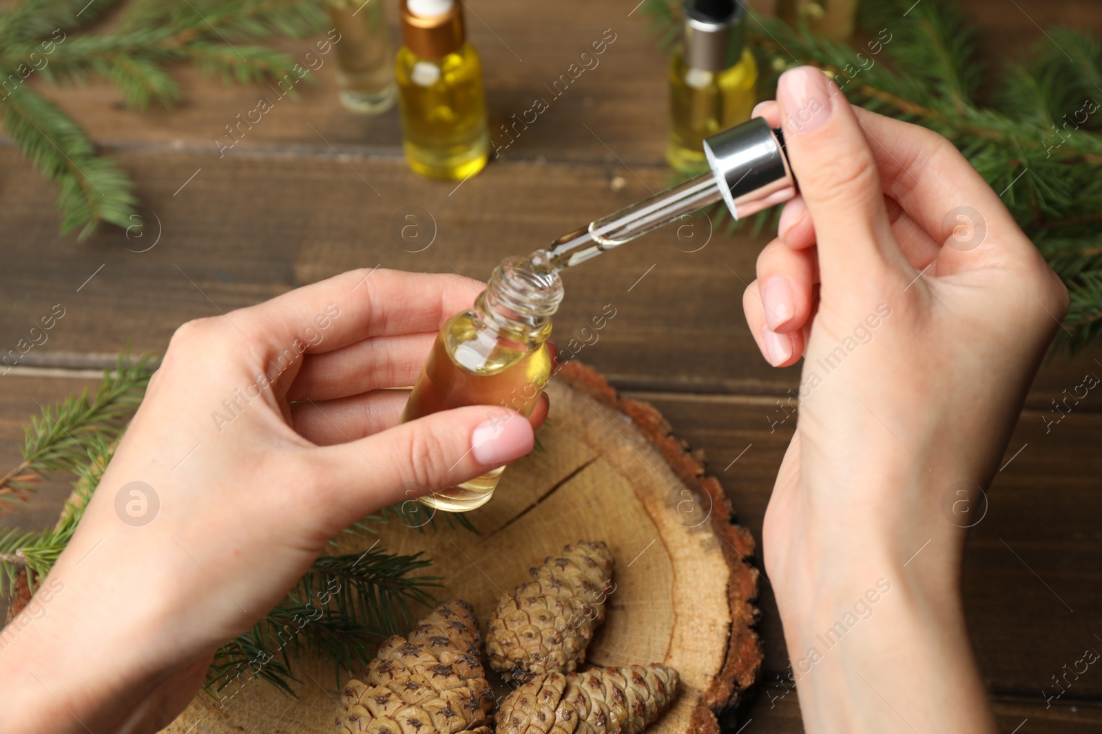 Photo of Woman holding pipette with pine essential oil over bottle at wooden table, closeup