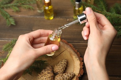 Photo of Woman holding pipette with pine essential oil over bottle at wooden table, closeup