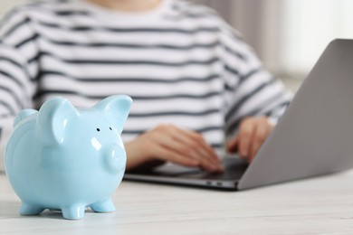 Financial savings. Woman using laptop at white wooden table indoors, focus on piggy bank