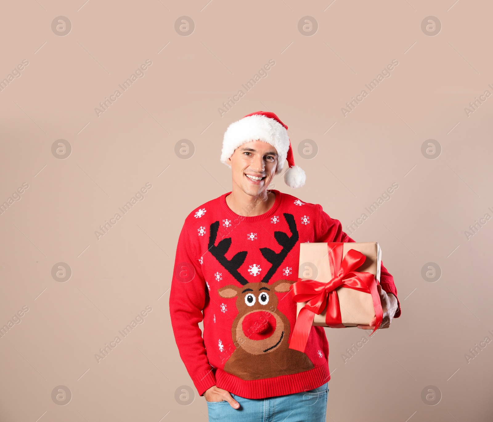 Photo of Happy man in Christmas sweater and Santa hat holding gift box on beige background