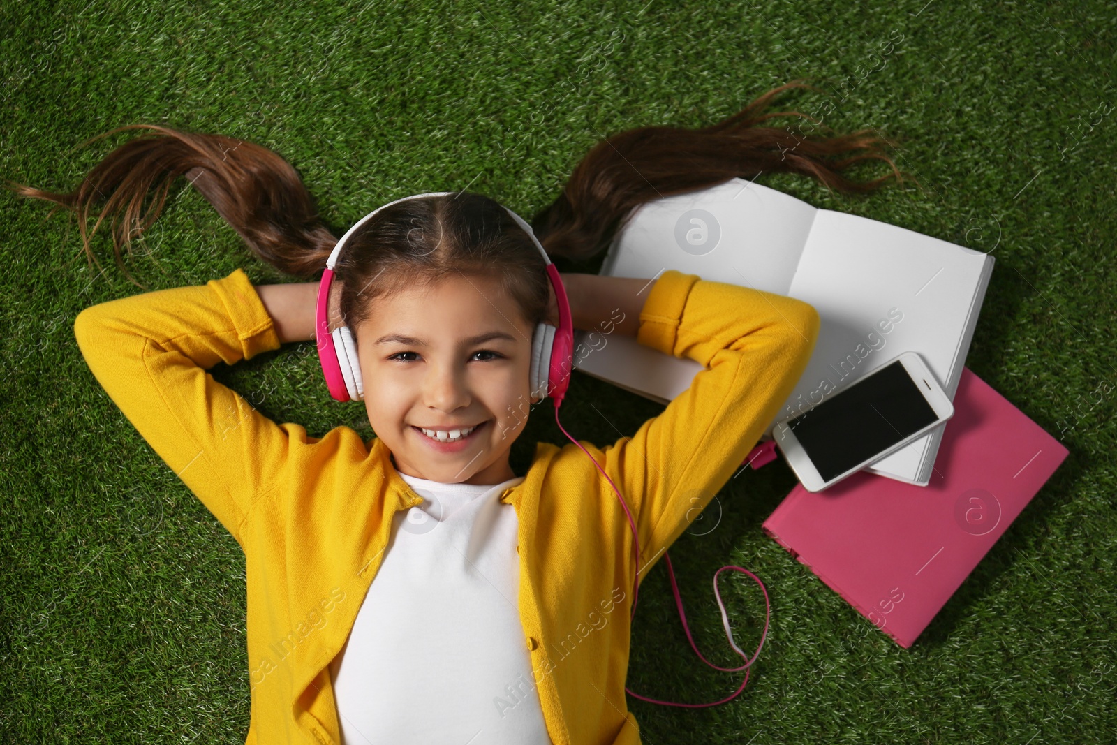 Photo of Cute little girl listening to audiobook on grass, top view