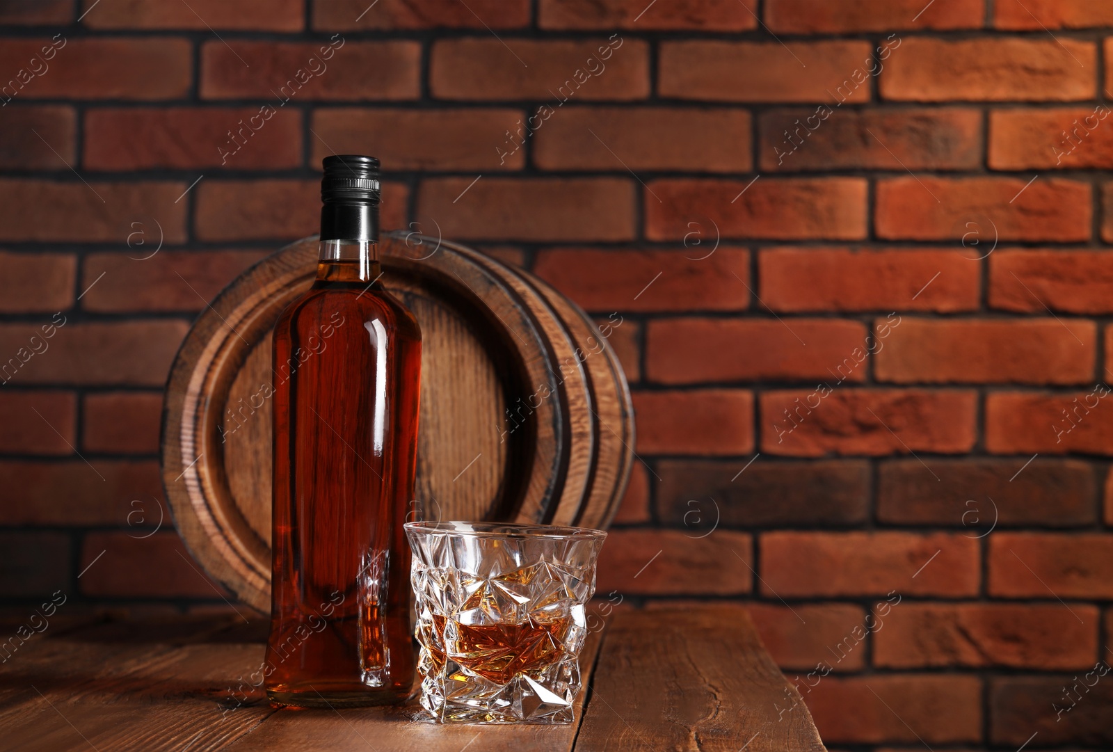 Photo of Glass and bottle of whiskey with wooden barrel on table against brick wall, space for text