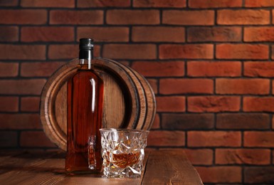 Glass and bottle of whiskey with wooden barrel on table against brick wall, space for text