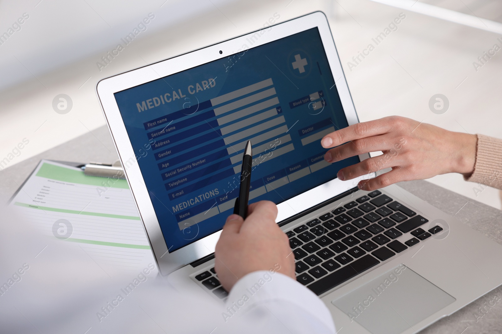 Photo of Doctor using laptop to fill out patient's medical card at table in clinic, closeup