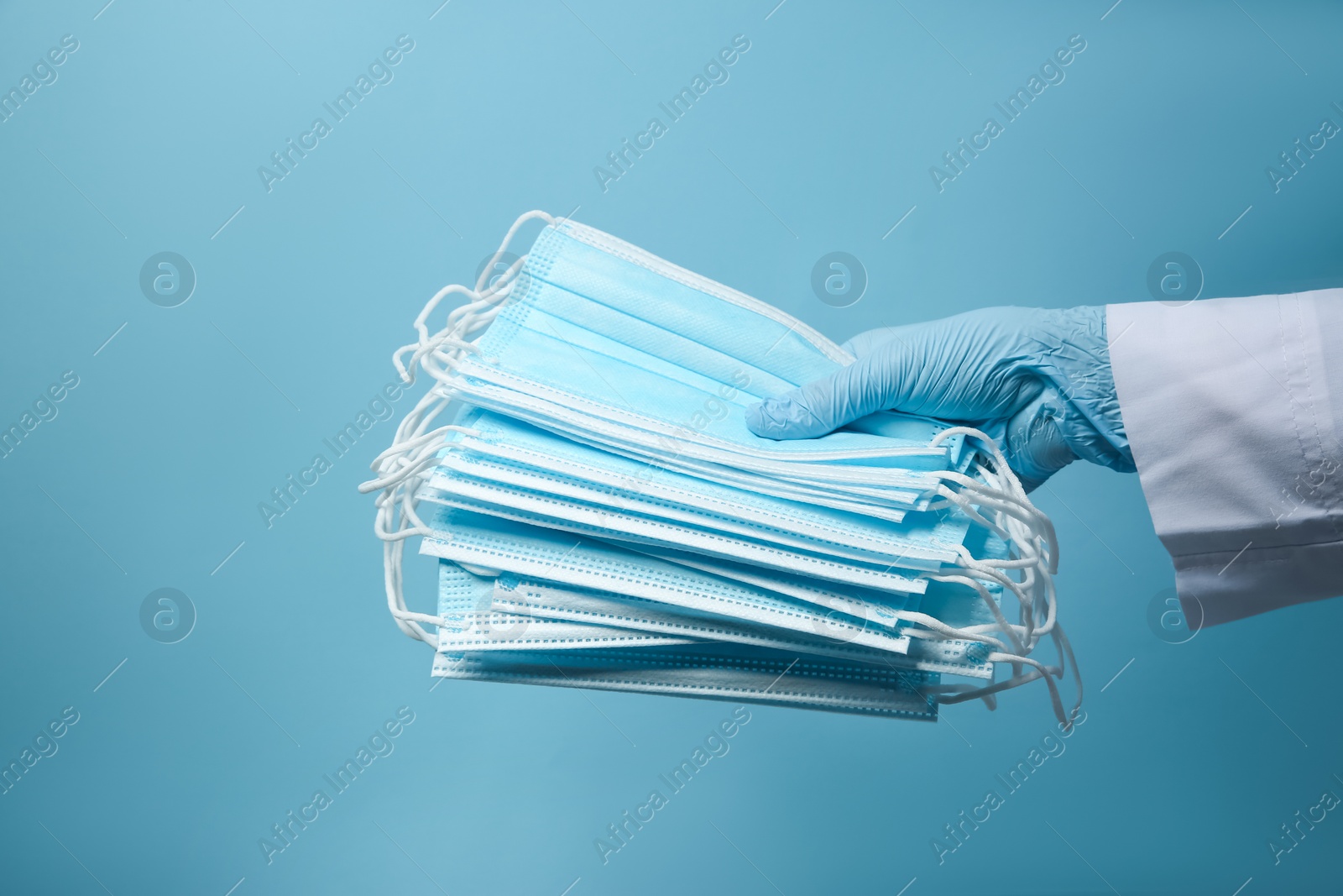 Photo of Doctor in latex gloves holding disposable face masks on light blue background, closeup. Protective measures during coronavirus quarantine