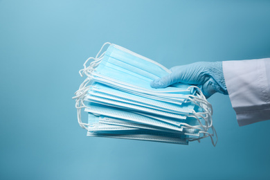 Doctor in latex gloves holding disposable face masks on light blue background, closeup. Protective measures during coronavirus quarantine