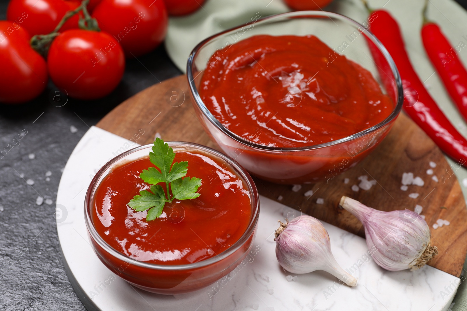 Photo of Organic ketchup in bowls and garlic on black table, closeup. Tomato sauce