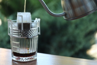 Photo of Pouring hot water into glass with drip coffee bag from kettle on wooden table, closeup