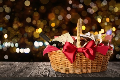 Image of Wicker basket full of gifts on wooden table against blurred festive lights. Space for text