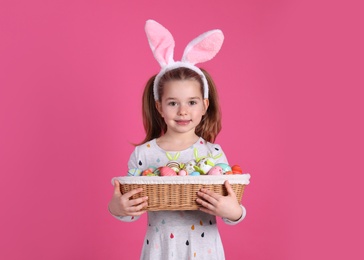 Photo of Adorable little girl with bunny ears holding wicker basket full of Easter eggs on pink background