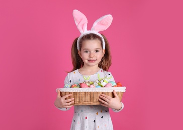 Adorable little girl with bunny ears holding wicker basket full of Easter eggs on pink background