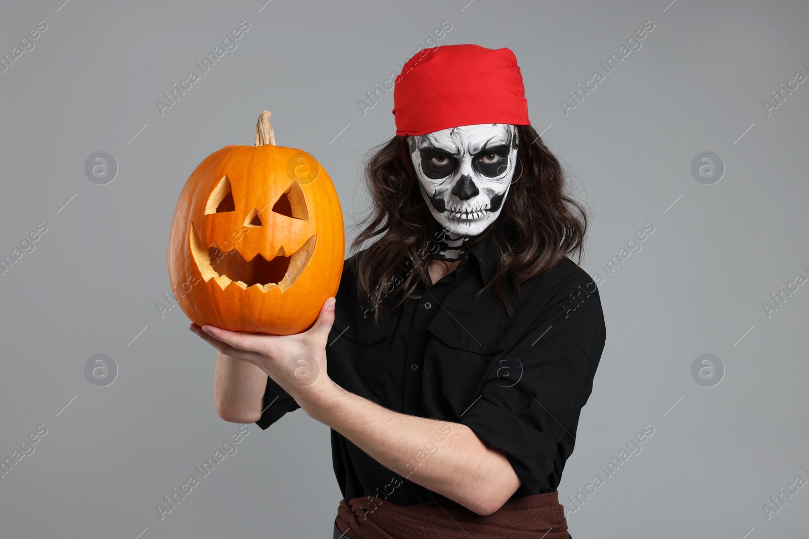 Photo of Man in scary pirate costume with skull makeup and carved pumpkin on light grey background. Halloween celebration