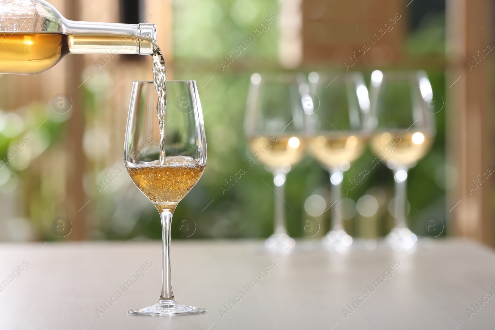 Photo of Pouring white wine from bottle into glass on table against blurred background, space for text