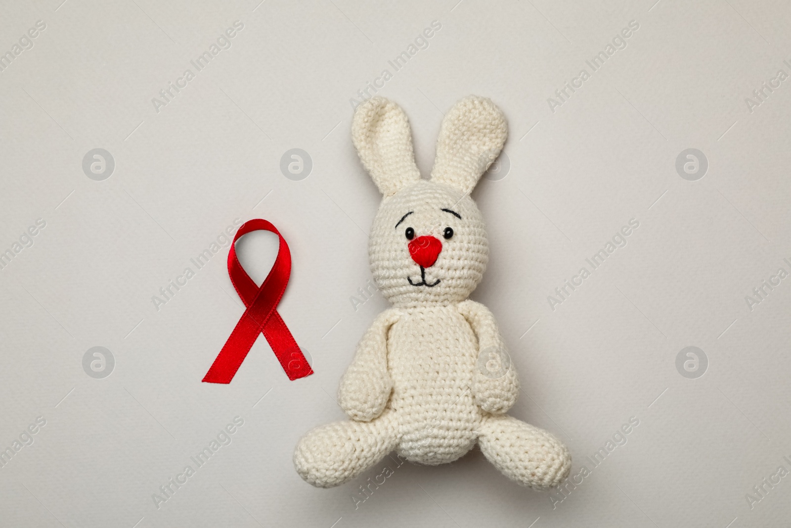 Photo of Cute knitted toy bunny and red ribbon on beige background, flat lay. AIDS disease awareness