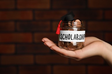 Woman holding glass jar of coins and graduation cap against brick background, closeup with space for text. Scholarship concept