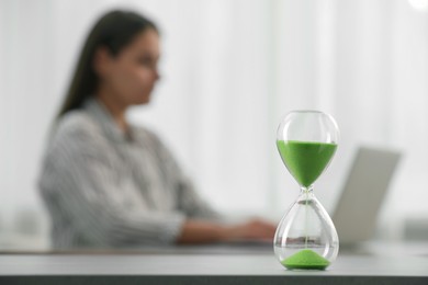 Photo of Hourglass with light green flowing sand on table. Woman using laptop indoors, selective focus