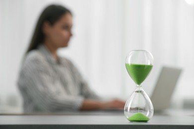 Hourglass with light green flowing sand on table. Woman using laptop indoors, selective focus