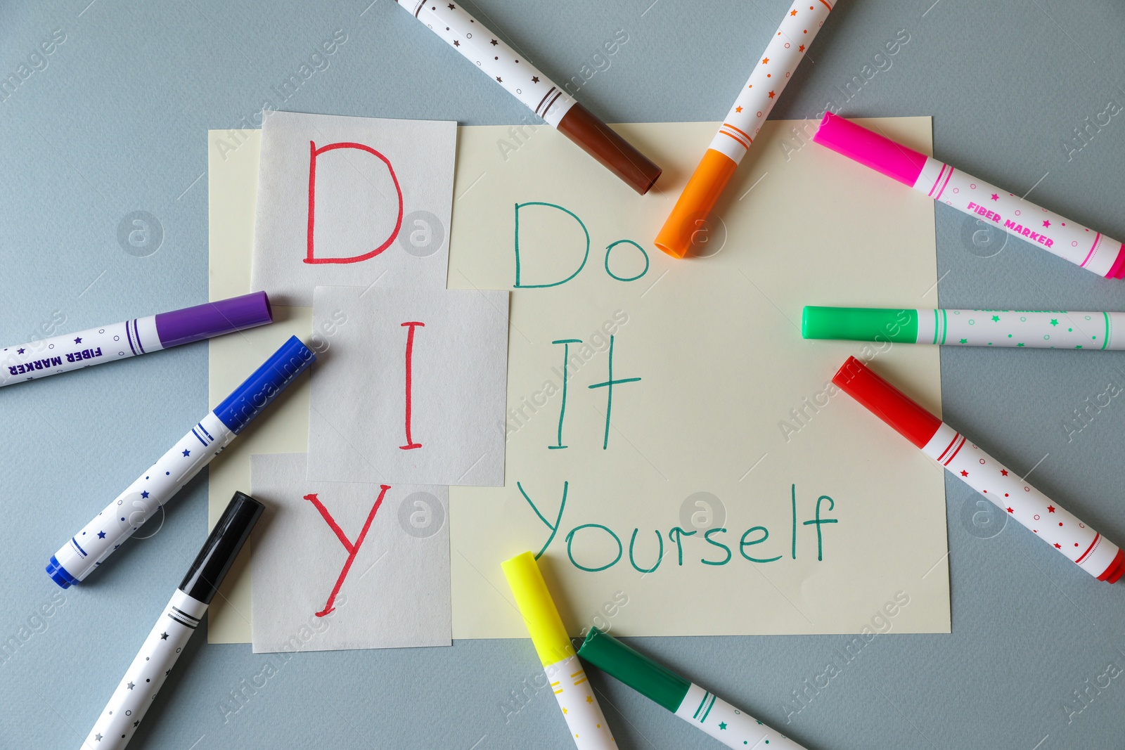 Photo of Sheet with written phrase Do It Yourself and colorful markers on grey background, flat lay. DIY concept