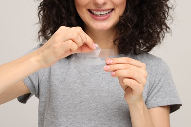 Photo of Young woman holding teeth whitening strips on light grey background, closeup