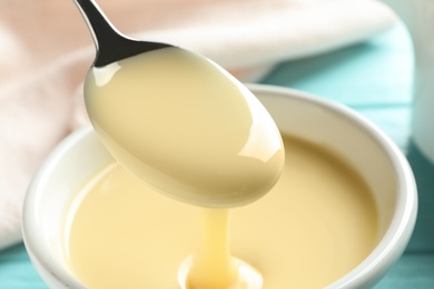 Photo of Spoon of pouring condensed milk over bowl on table, closeup. Dairy products